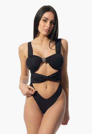Bikini nero con design frontale attorcigliato, spalline larghe e un elemento decorativo in metallo argentato, caratterizzato da un fondo a taglio alto. Morbida al tatto.