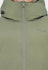 Giacca imbottita verde con colletto alto, dotata di una cerniera frontale, texture liscia e logo "REPLAY" stampato di lato.