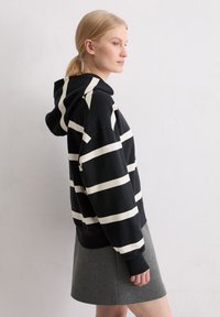 Black pullover hoodie with wide white stripes, featuring a drawstring hood, ribbed cuffs, and hem. Paired with a grey skirt.