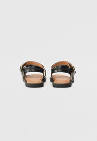 Sorte lær sandaler med en brun såle, med justerbar rem og en liten gullspenne. Åpen bakdesign for pusteevne.