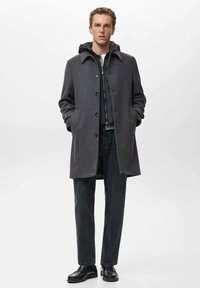 Abrigo gris de lana con cierre de botones, que cuenta con una capucha. Combinado con jeans oscuros y zapatos negros. Diseño simple, con bolsillos prácticos.