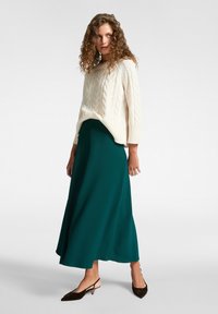 Knit-Creme-Pullover mit Zopfmuster, kombiniert mit einem fließenden teal-farbenen Midikleid und dunklen, spitzen Slingback-Schuhen.