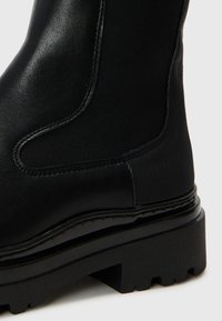 Botim preto até ao tornozelo feito de couro liso, com painéis laterais elásticos, bico arredondado e sola de borracha texturizada com um sulco profundo.