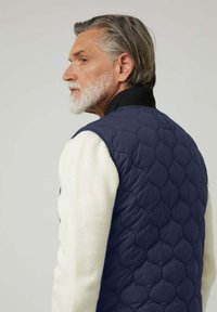 Chaleco acolchado azul marino con cuello alto, combinado con un suéter crema. El chaleco presenta un patrón texturizado y es sin mangas.