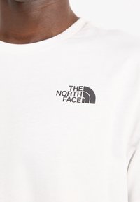 Vit bomullströja med rund halsringning och korta ärmar, med en svart logotyp av "The North Face" på bröstet. Slät textur.