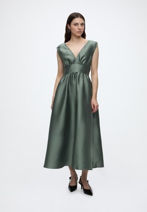 Femme en robe sans manches en satin vert foncé avec un décolleté en V et une taille froncée, portant des talons hauts noirs à bout pointu, debout sur un fond blanc.