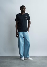 Mees seisab, seljas must graafilise mustriga t-särk, lahtised helesinised teksad ja valged tossud, taustal helehall sein.