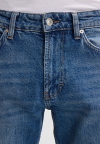 Jeans in denim blu con chiusura a bottoni, caratterizzati da cuciture a contrasto e design classico a cinque tasche. La texture appare leggermente robusta.