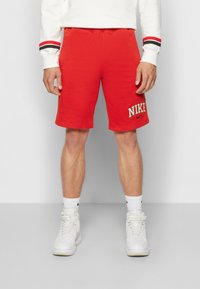 Červené bavlněné šortky s elastickým pasem, s vyšitým bílým logem "NIKE" na levé noze, doplněné bílými teniskami.