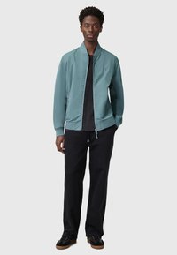 Giacca bomber in teal chiaro con zip frontale, polsini a coste e tasche laterali, abbinata a pantaloni neri e scarpe scure. Tessuto di superficie liscio.