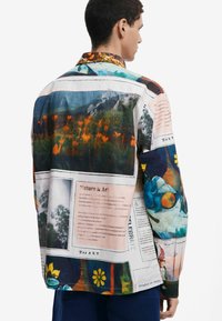 Camicia button-up con una stampa mista di paesaggi e motivi floreali in colori vivaci, con una finitura lucida e un colletto a punta.