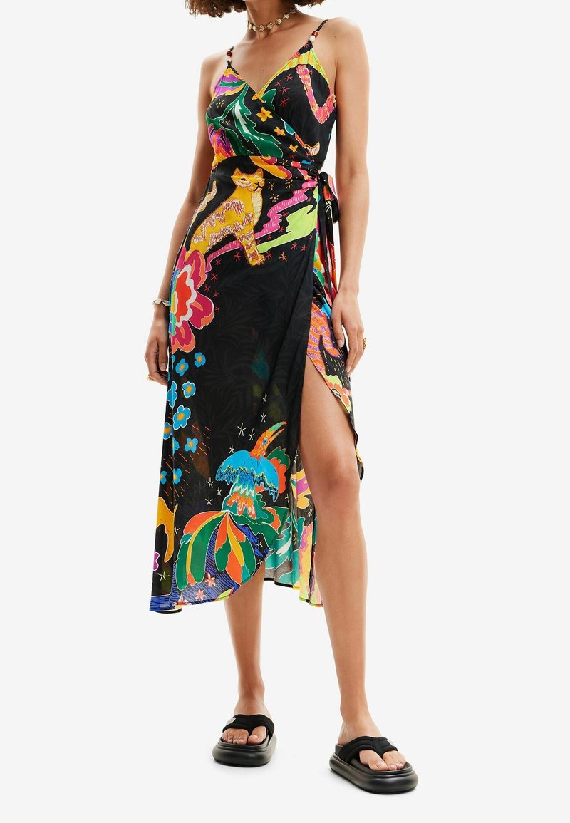 Femme portant une robe portefeuille colorée avec des motifs floraux et animaliers, une fente haute sur la cuisse, des fines bretelles et des sandales compensées noires.