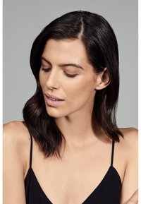 Haut à fines bretelles noires en tissu doux ; mannequin affichant des cheveux bruns foncés lisses et un maquillage neutre. Textures subtiles visibles sur la peau.