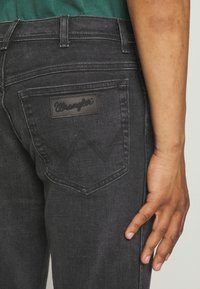 Juodi džinsai su galiniu kišeniu ir stačiakampiu juodu odos ženkliuku su užrašu "Wrangler" bei išskirtinėmis siūlėmis, išryškinančiomis kišenes.