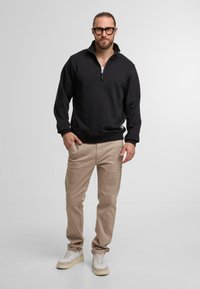 Sudadera nera con zip e colletto a coste, abbinata a pantaloni beige e sneakers chiari. L'uomo è in piedi su uno sfondo neutro.