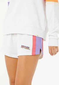 Shorts blancs avec des rayures latérales roses et violettes, arborant un logo "RIPCURL". Le tissu est doux et le design comprend des fentes sur les côtés.