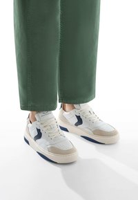Sneaker bianchi e beige con superficie testurizzata, dettagli in blu navy e perforazioni. Indossati con pantaloni cropped verdi.