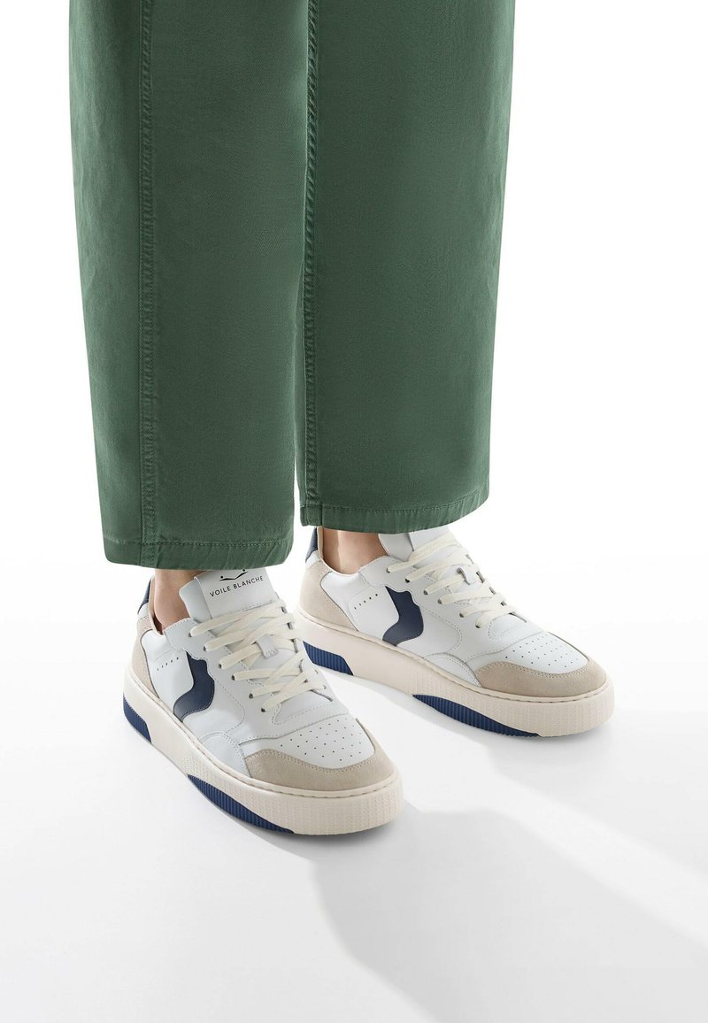 Sneaker bianchi e beige con superficie testurizzata, dettagli in blu navy e perforazioni. Indossati con pantaloni cropped verdi.