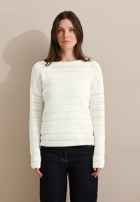 Pull en tricot blanc avec un motif texturé et rayé, un col rond et des manches longues ; associé à un jean en denim foncé.
