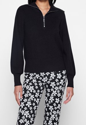 Femme portant un pull noir à côtes avec une fermeture éclair à l'avant et un pantalon noir avec un motif floral blanc.