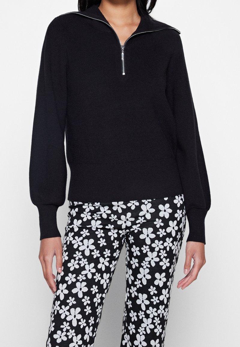 Femme portant un pull noir à côtes avec une fermeture éclair à l'avant et un pantalon noir avec un motif floral blanc.