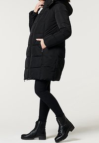 Černá péřová bunda s kapucí, obsahující zipový uzávěr, prošívaný vzor a boční kapsy. V kombinaci s černými skinny kalhotami a botami.