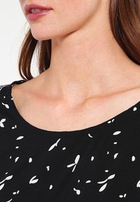 Haut en tissu noir avec un décolleté arrondi et un motif blanc en forme de feuilles. Texture lisse avec des manches courtes, présentant un design moderne.