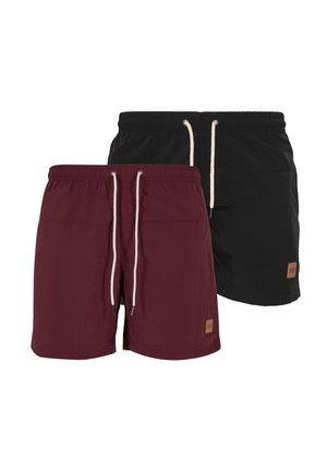 Homme portant un short bordeaux à cordon, un t-shirt blanc et des sandales noires à enfiler, debout devant un fond uni.
