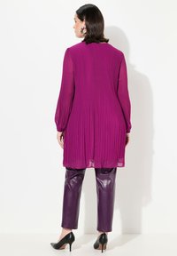 Magenta geplooide tuniek met lange mouwen, gecombineerd met glanzende paarse leren broek en zwarte stilettohakken. Achteraanzicht weergegeven.