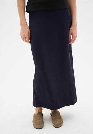 Jupe navy longueur cheville en tissu lisse et extensible. Elle présente une taille ajustée et une silhouette légèrement évasée. Portée avec des chaussons beiges.