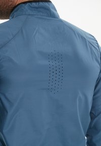 Blå vandtæt jakke med høj krave, der har et perforeret design detalje på ryggen for ventilation. Glat, letvægtsstof.