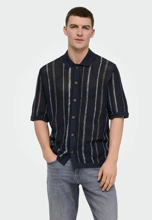 Giovane uomo che indossa una camicia blu navy a maniche corte con bottoni e righe verticali e jeans grigio chiaro, in piedi con una mano in tasca.