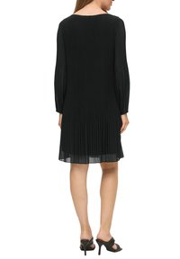 Schwarzes plissiertes Kleid mit langen Ärmeln, rundem Halsauschnitt und leichtem Stoff, abgerundet mit einem fließenden Saum. Getragen mit schwarzen High Heels.