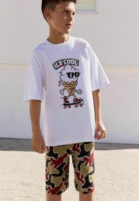 Witte T-shirt met een ijsje grafiek en een zonnebril; korte camouflagedragen shorts in groen, zwart en rood. Casual zomerset.