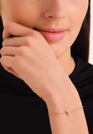 Pulsera de cadena de oro con un pequeño charm de cruz adornado con piedras transparentes, mostrado en una mano descansando cerca de la cara.