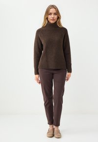 Bruine gebreide trui met hoge kraag, gecombineerd met donkergroene broek en lichtbruine instappers. Zachte textuur en een comfortabele pasvorm.