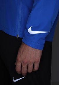 Veste de sport bleue à texture lisse, arborant un logo Nike blanc en forme de swoosh sur la manche. Fermeture éclair à l'avant et pantalon noir visible en dessous.
