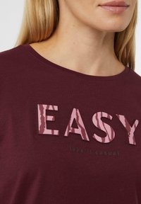 Bordeauxfarbenes T-Shirt mit Rundhalsausschnitt, das mit strukturiertem pinken Schriftzug "EASY" und einem kleineren Text "take it casual" darunter versehen ist. Weiche Textur des Stoffes.