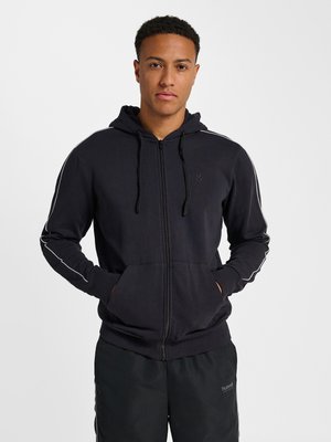 Ung mand iført en sort hoodie med lynlås og hvide striber på ærmerne og matchende sorte bukser, stående med hænderne i hoodie-lommerne.