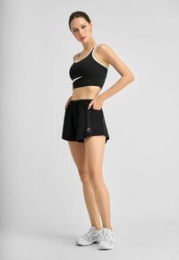 Schwarzes sportliches Crop-Top mit weißem Rand, kombiniert mit schwarzen Shorts mit Kordelzug und Seitentaschen, getragen mit weißen Sneakers.