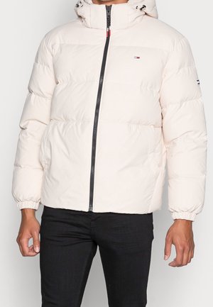 Man draagt een roomkleurige donzen jas met capuchon, zwarte rits en kleine Tommy Hilfiger-logo's op de borst en mouw.