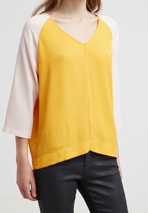 Kobieta ubrana w luźną bluzkę z musztardowożółtą górą i jasnoróżowymi rękawami do trzy czwarte długości, w połączeniu z czarnymi spodniami.