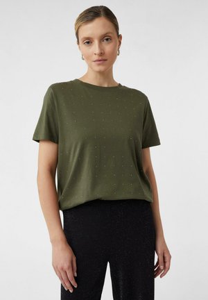 Frau mit nach hinten gegeltem Haar, die silberne Ohrreifen, ein olivgrünes, mit Nieten besetztes, kurzärmliges Oberteil und schwarze, glitzernde Hosen trägt, steht vor einem weißen Hintergrund.