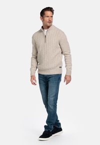 Beige ribbeltrui met een half-rits kraag, gedragen met blauwe jeans en zwarte schoenen. De trui heeft een gestructureerd breipatroon.