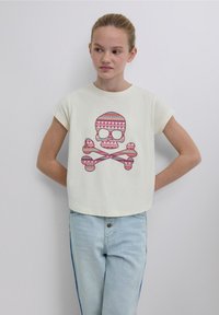 Cremefarbenes T-Shirt mit kurzen Ärmeln und einem großen grafischen Aufdruck eines Schädels mit rosa und türkisfarbenen Mustern, kombiniert mit hellblauen Jeans.