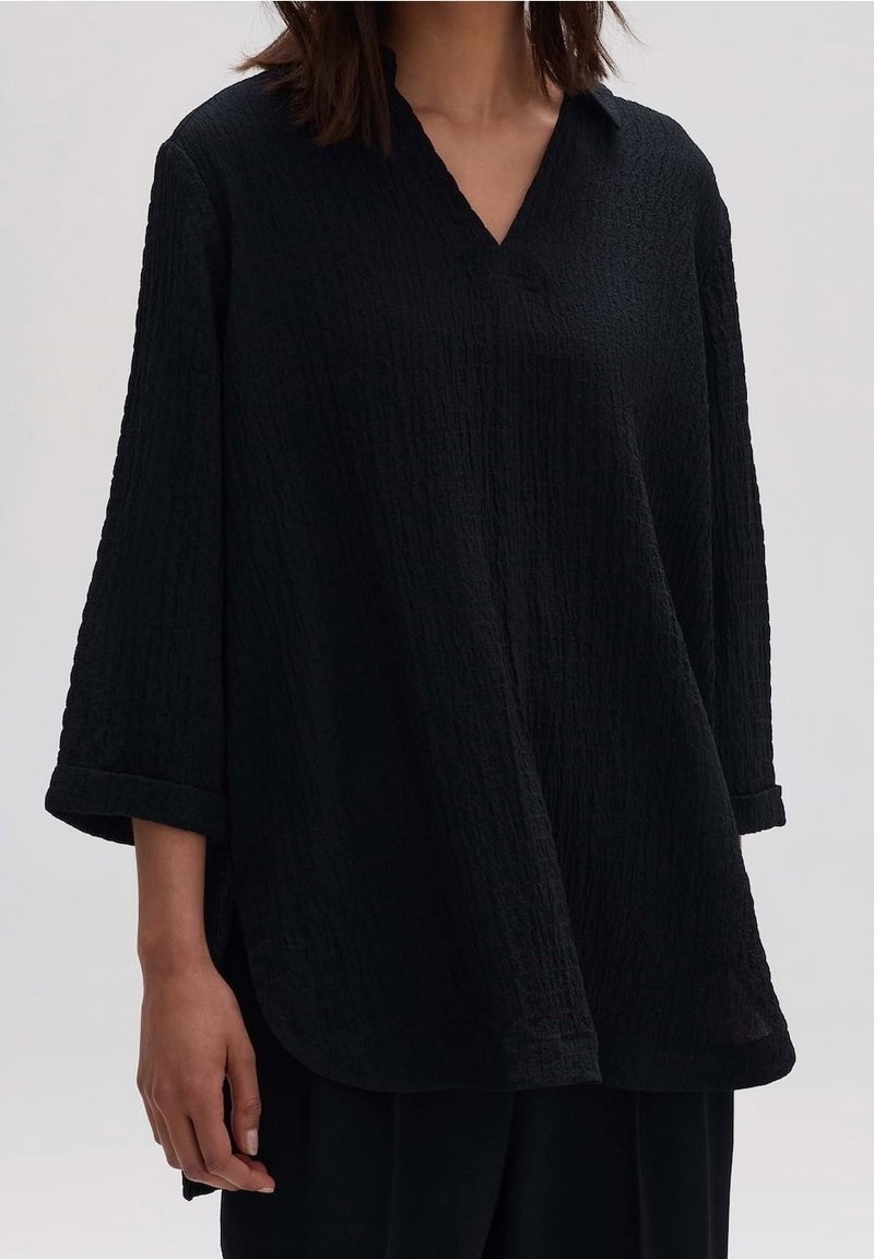 Blouse oversized noire texturée avec un décolleté en V et des manches trois-quarts, présentant un motif légèrement froissé et des fentes sur les côtés.