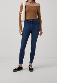 Tmavě modré skinny džíny s vysokým pasem, nošené s světle hnědým topem s odhalenými rameny a černými teniskami s bílými tkaničkami.