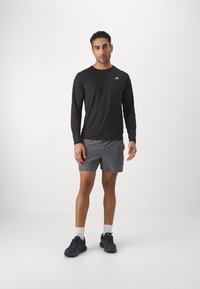 Camisa preta de manga longa com um pequeno logótipo, acompanhada de calções cinzentos. Usada com sapatilhas desportivas escuras e meias brancas. Design simples.