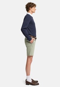 Marineblå langærmet skjorte over en hvid krave-skjorte, parret med lysegrønne shorts. Brun bælte og loafers med hvide sokker synlige.
