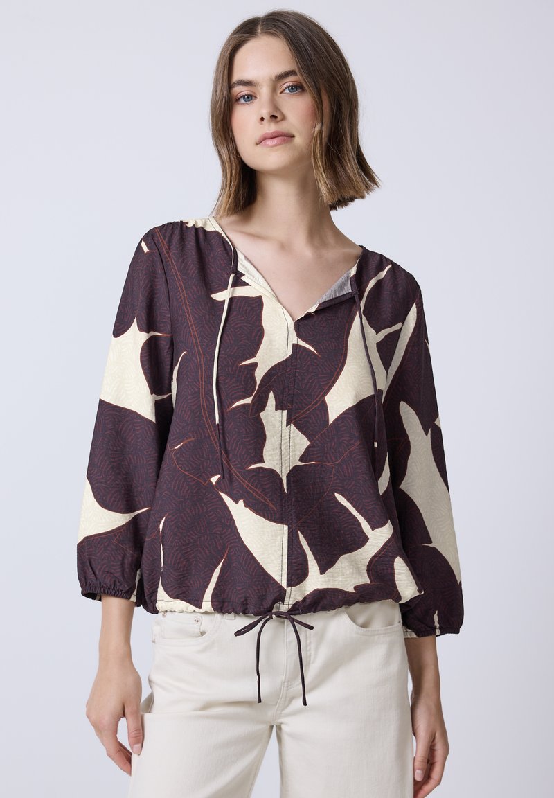 Blouse à capuche dans un motif de feuilles bordeaux foncé et crème. Présente des poignets élastiques et une taille à cordon. Tissu léger avec un design texturé.